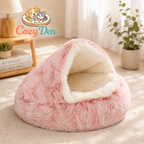 A pink plush pet bed with a cream-colored interior, styled in a warm, sunlit room with a wooden side table, lamp, and potted plant. The CozyDen brand logo is visible in the upper left corner.