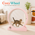 A pink cat exercise wheel with a tabby cat walking inside, set in a bright, modern living room. The brand name 'Cozy Wheel' is displayed at the top left.