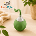 A green CozyRoller pet toy or accessory with a curved green handle and a clear plastic neck, shown on a beige tabletop with a knit blanket, potted plant, and the CozyRoller logo in the upper left.